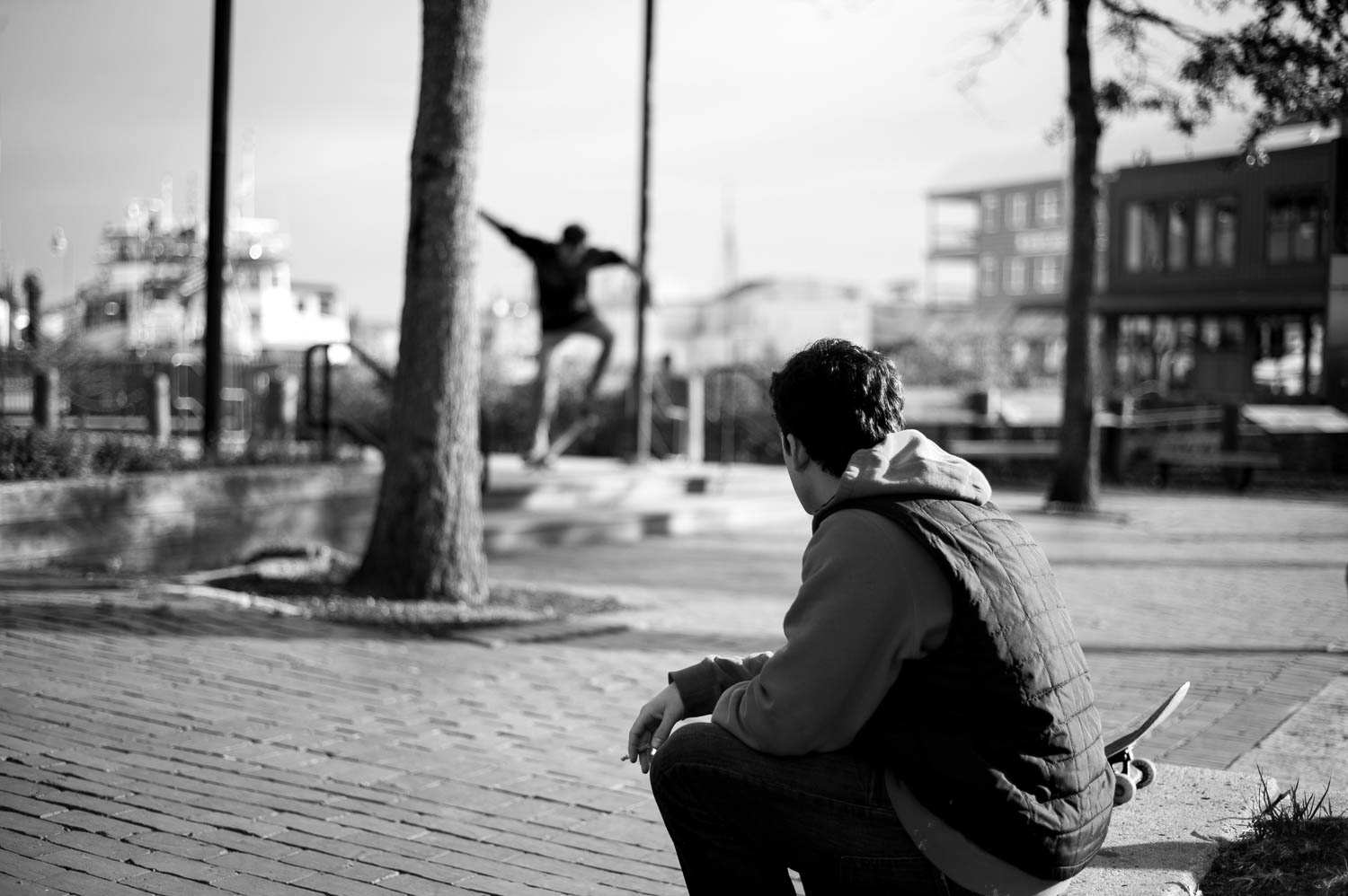 A skateboarder smoking a cigarette with his friend skateboarding in the background behind him