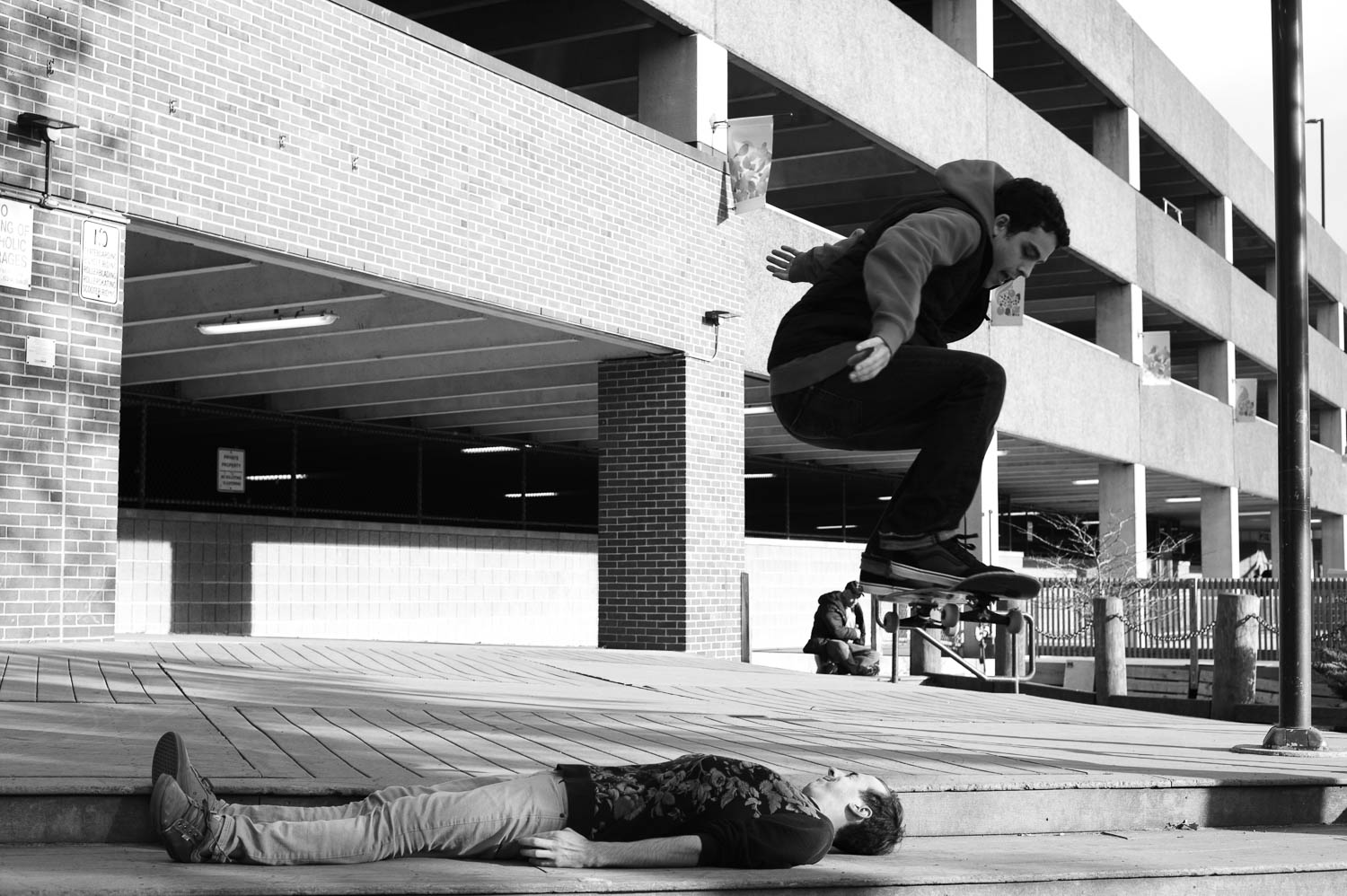 A skateboarder suspended in air above his friend, who is lying down and squinting