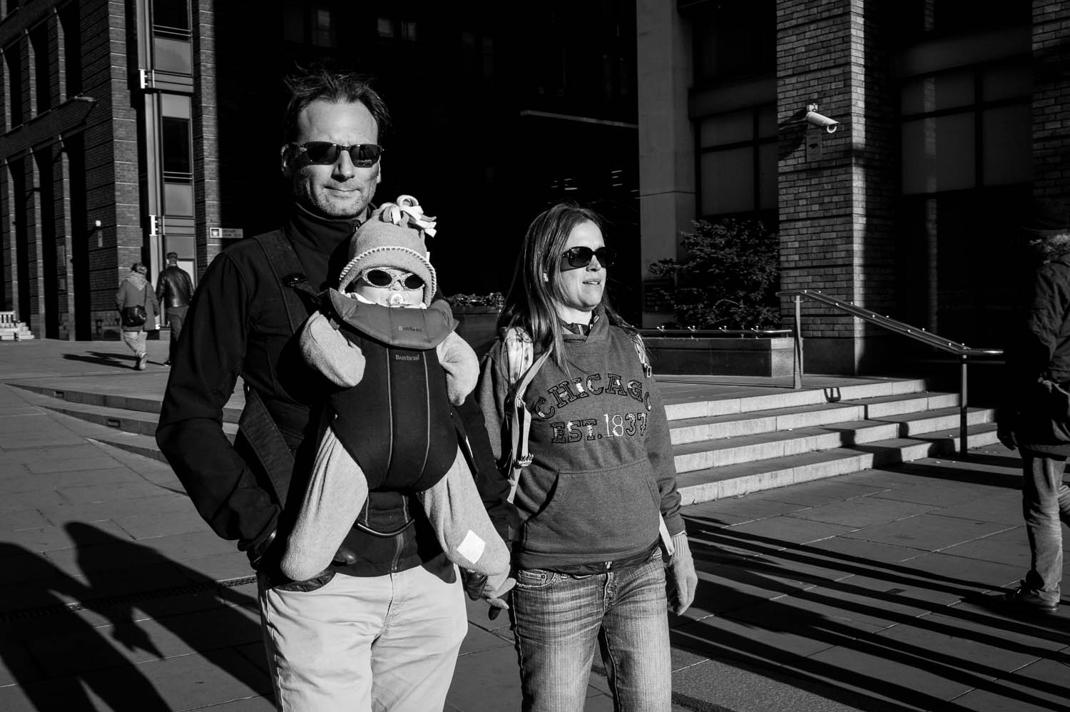 A couple with a baby carrier walk through London, all three wearing sunglasses