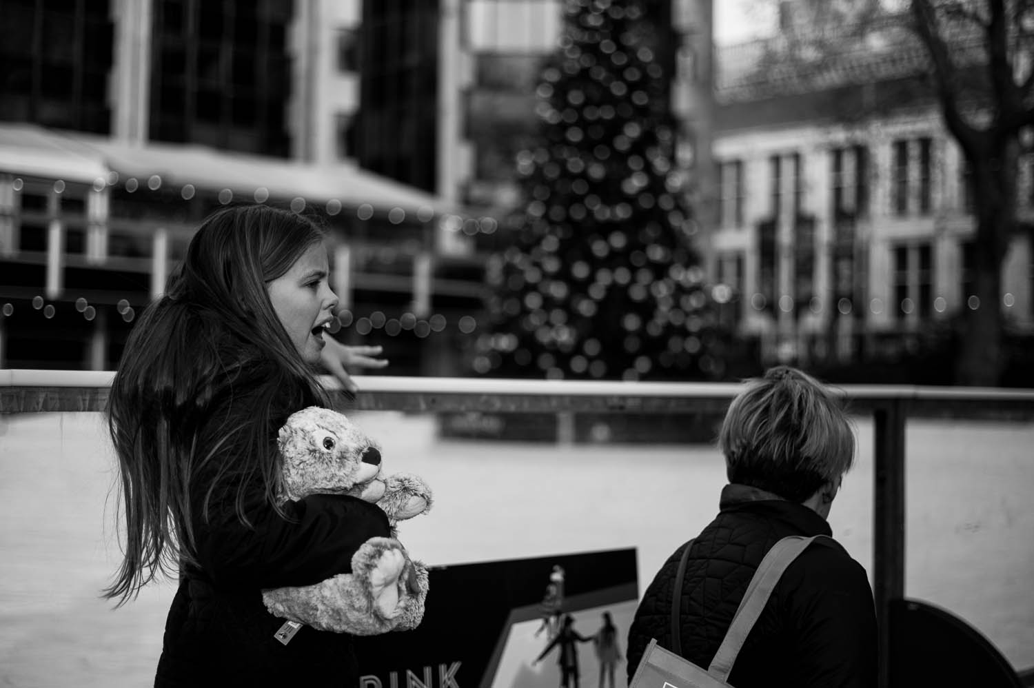 A small girl holding a stuffed animal in front of an outside ice rink and Christmas tree in London