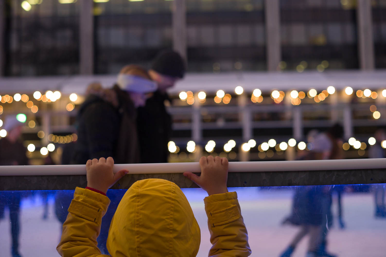 A child in a yellow jacket peers over the rail of an outside skating rink in London