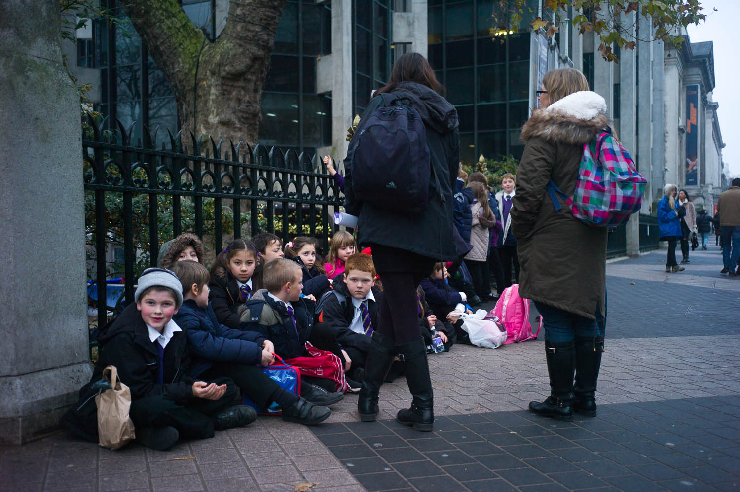 Schoolchildren are seated against a fence near the museums in London