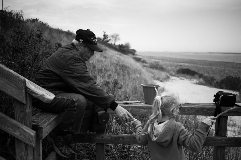 A man in a Korea Veteran hat holds the hand of a young girl playing on the beach