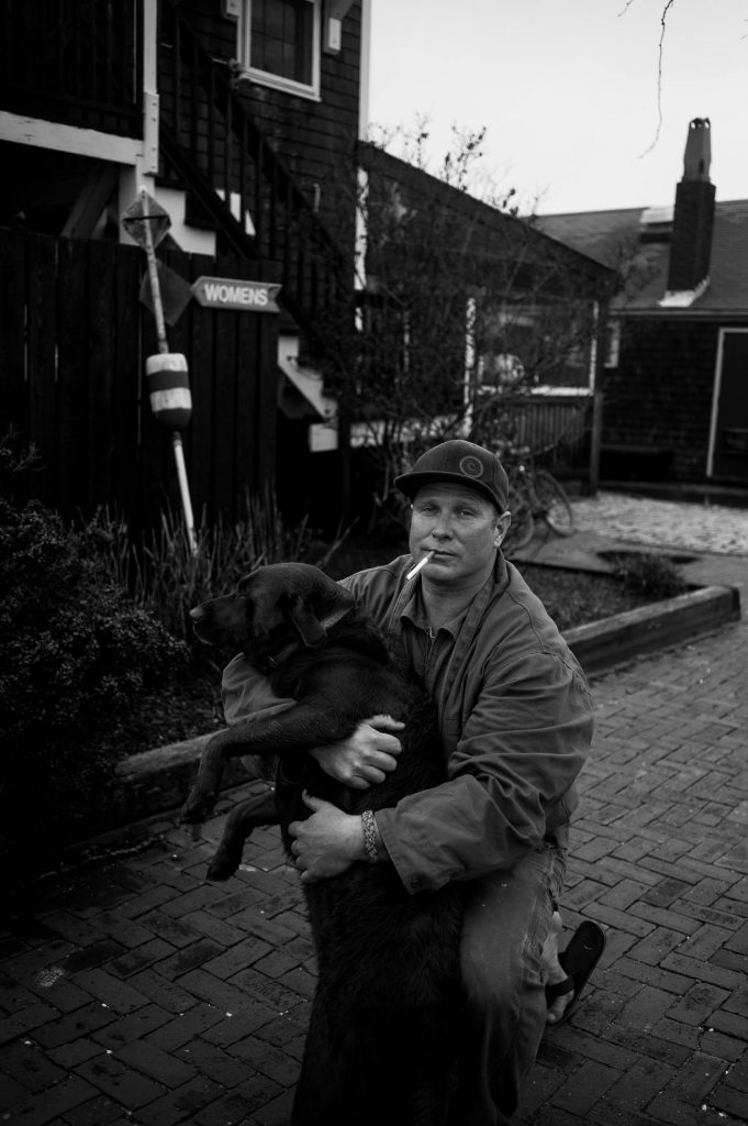 A man with a cigarette in his lips holding his dog for a portrait.