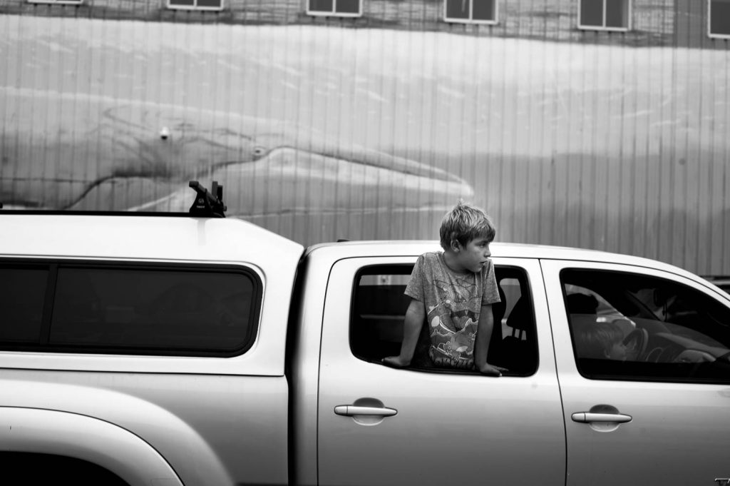 A young boy with a Pokémon Charizard evolution shirt leaning out the back passenger-side window of a pickup truck with closed cab on its way onto the ferry to Peaks Island